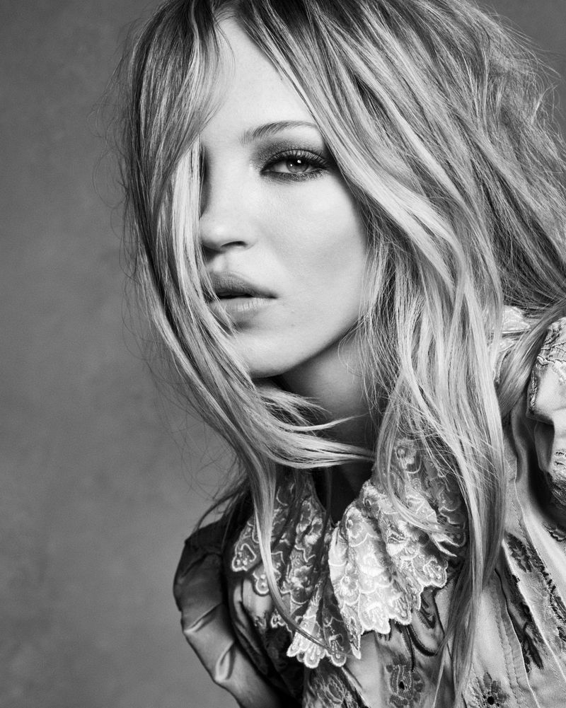 Black and white close-up portrait of model Lila Moss with loose blonde hair falling across her face against a plain grey background.