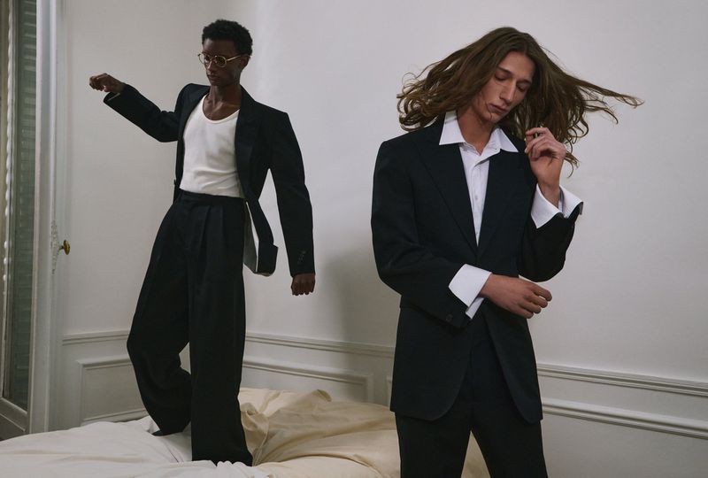 Two male models in dark suits posing indoors against a white wall with wainscoting, captured for an editorial project by Reto Schmid.