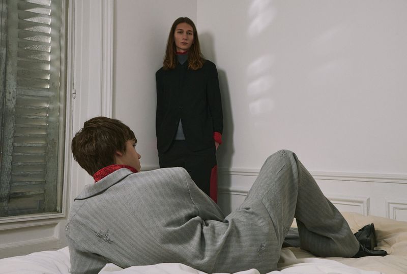 Two people pose indoors: One adult with long hair stands by a wall while another sits on a bed wearing a grey suit.