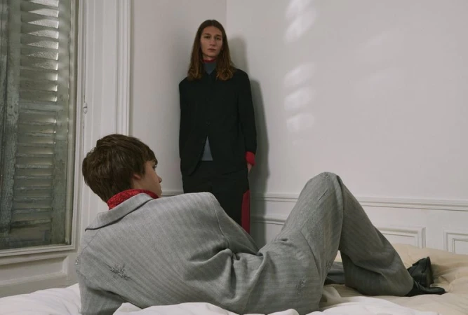 Two people pose indoors: One adult with long hair stands by a wall while another sits on a bed wearing a grey suit.