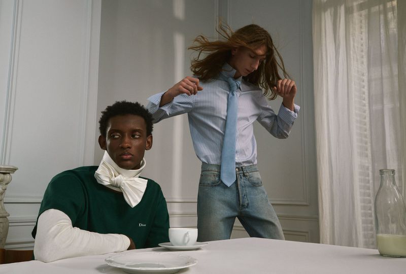 Two male models posing indoors with a seated man in a green shirt and a standing man with long hair in a striped shirt