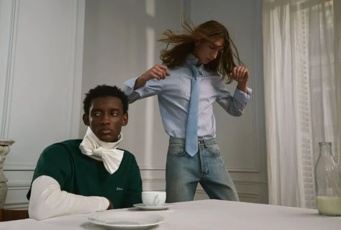 Two male models posing indoors with a seated man in a green shirt and a standing man with long hair in a striped shirt