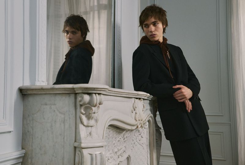 Young man in a dark blazer leaning against a white marble mantelpiece with a mirror reflection
