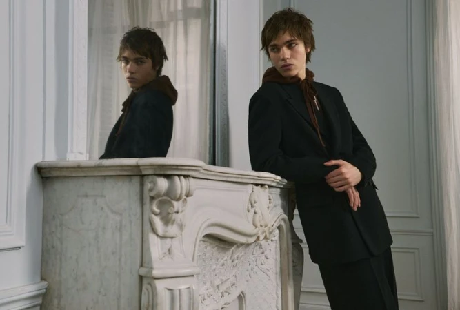 Young man in a dark blazer leaning against a white marble mantelpiece with a mirror reflection