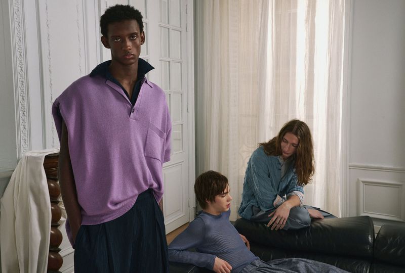 Three young men posing in an interior space with white paneled walls, with one standing hero model wearing a purple knit polo.