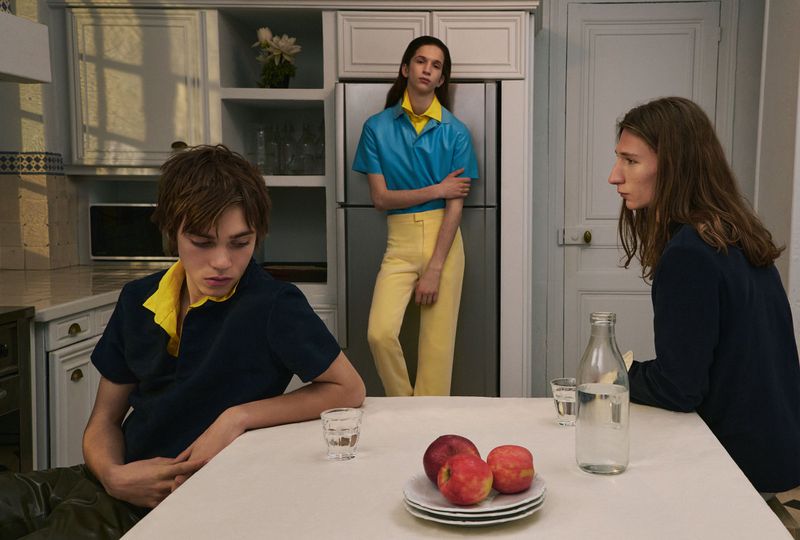 Three models in a kitchen setting with a table holding a plate of red apples and a glass water bottle.