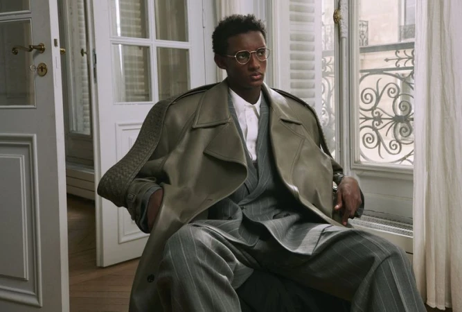 Model wearing eyeglasses and an oversized coat seated inside a room near tall windows