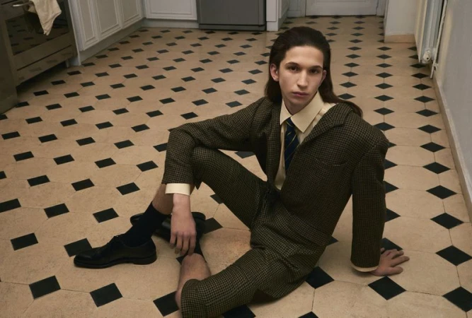 Model with long dark hair wearing a patterned green suit seated on a diamond-patterned tiled floor in a kitchen