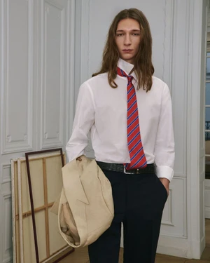 Model with long brown hair wearing a white shirt and striped tie, holding a beige jacket in an interior setting