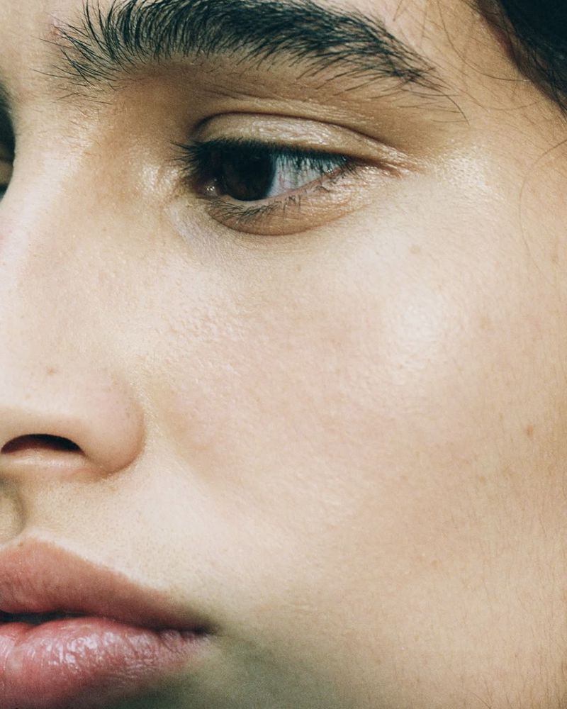 Close-up studio shot of a young adult female's face featuring her brown eye, eyebrow, skin texture, and full lips, with soft, neutral lighting.