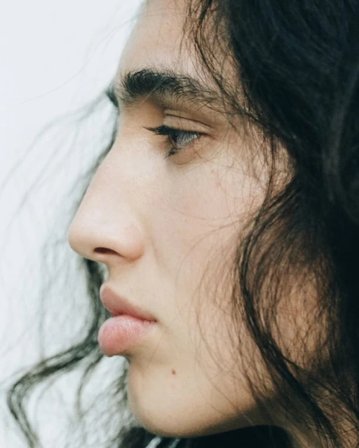 Close-up portrait of a person with wavy dark hair in profile against a white background, showcasing facial features and hair texture.
