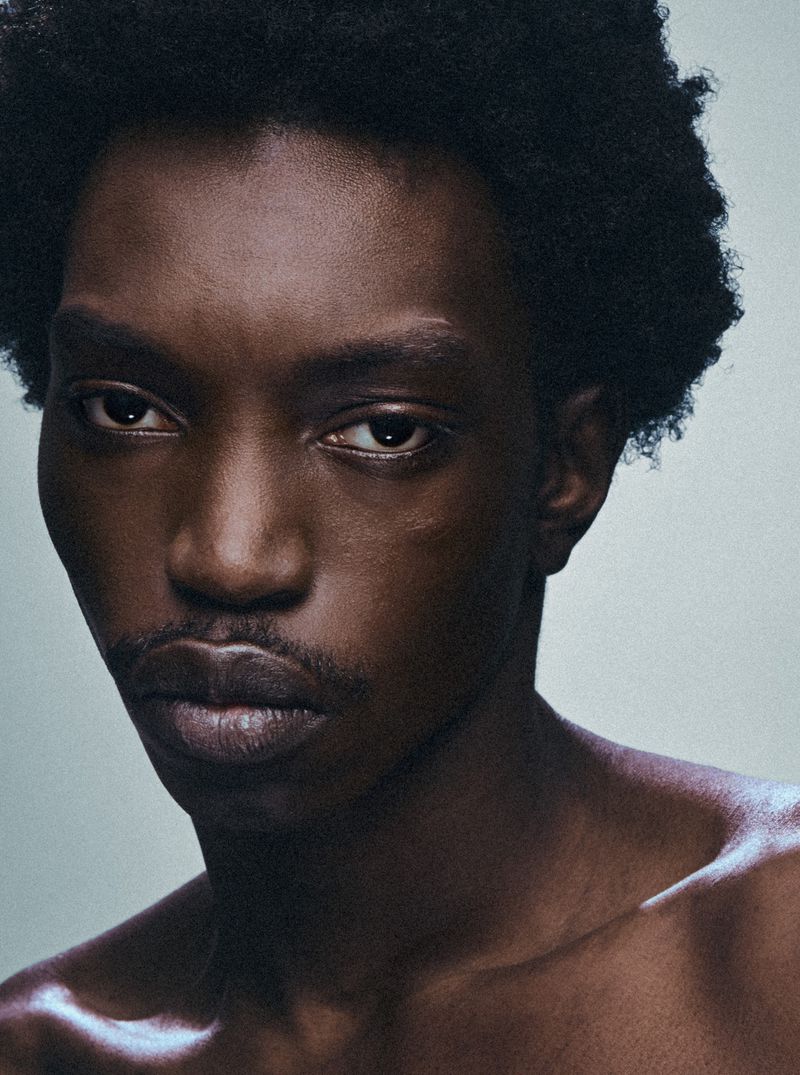 Close-up portrait of a young Black man with dark curly hair against a plain light grey backdrop for HERO USA.