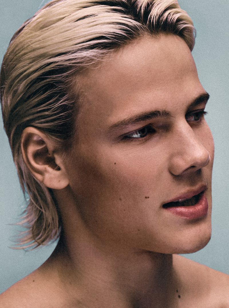 Close-up portrait of a young adult male with blond hair, fair skin, and neutral expression against a solid light blue background.