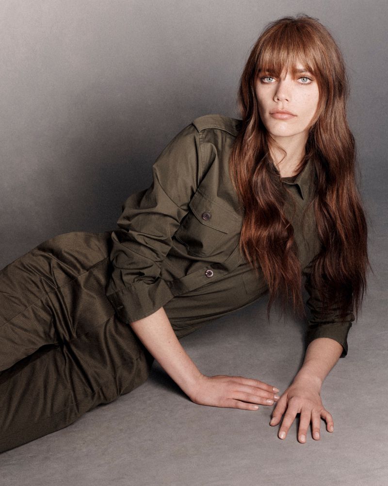 Woman with long brown hair and bangs wearing an olive-green jumpsuit lying down in a photo studio with a grey background.