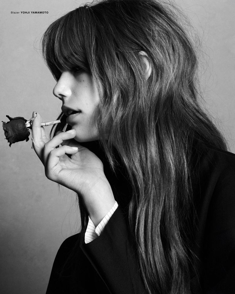 Profile portrait of a woman holding a dark rose near her lips, wearing a black blazer and pinstripe shirt.