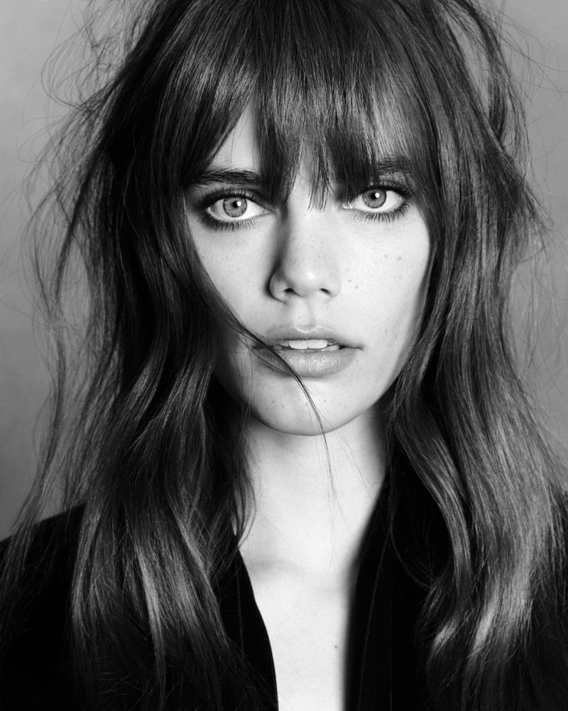 Close-up portrait of a woman with long dark hair and bangs, wearing dark clothing against a plain background.