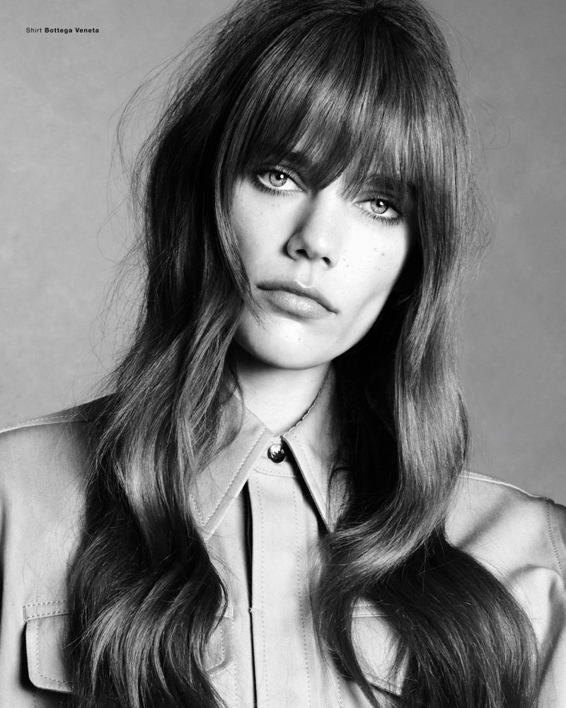 Close-up monochrome portrait of a model with long hair, full bangs, and a collared shirt against a grey background.