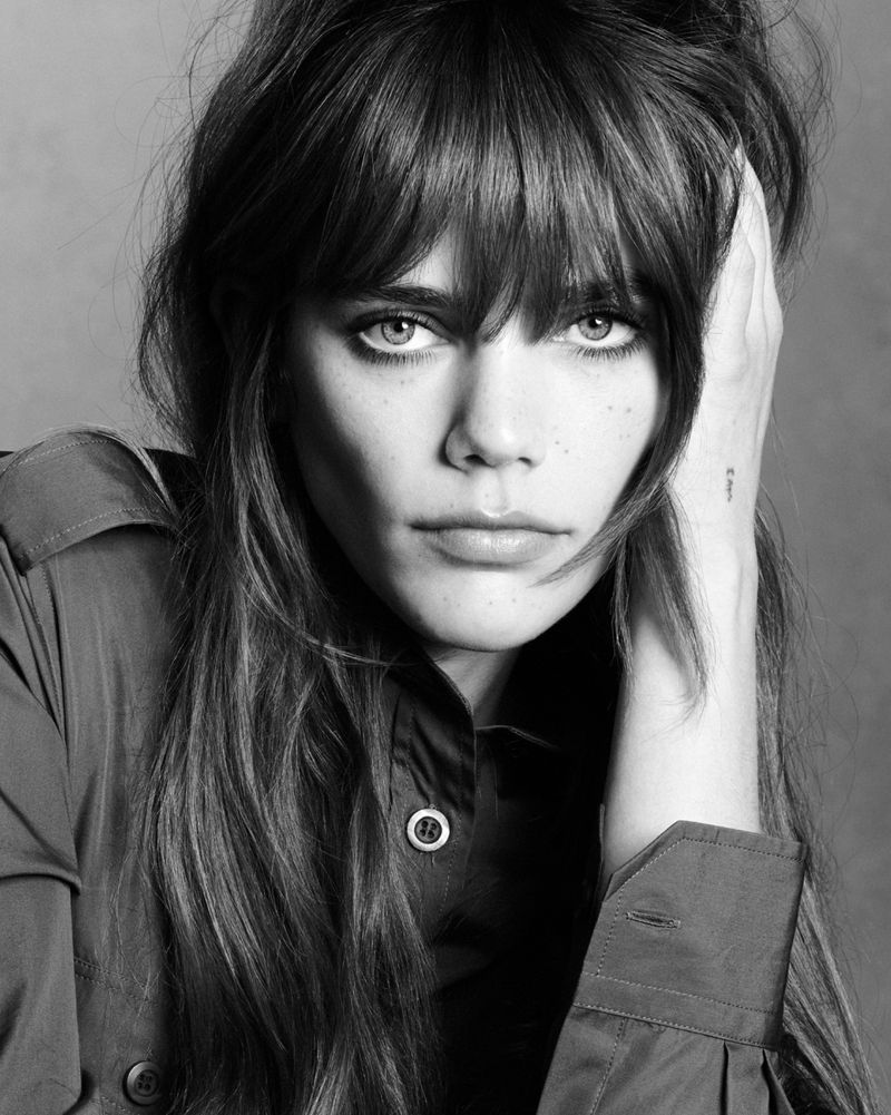 Close-up monochrome portrait of a woman with bangs wearing a button-down shirt, touching her hair against a plain studio background.