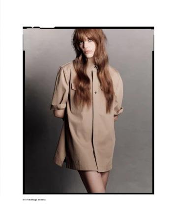 Model with long brown hair wearing a tan button-up shirt standing against a neutral grey studio background.