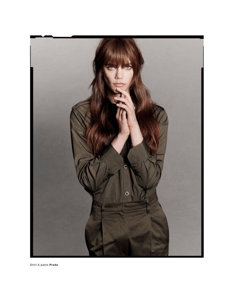 Model Betsy Gaghan wearing an olive green button-up shirt and trousers against a neutral grey studio background.