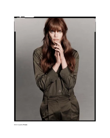 Model Betsy Gaghan wearing an olive green button-up shirt and trousers against a neutral grey studio background.