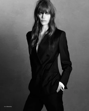 Model wearing a black tailored pantsuit with a white shirt cuff against a textured dark studio background