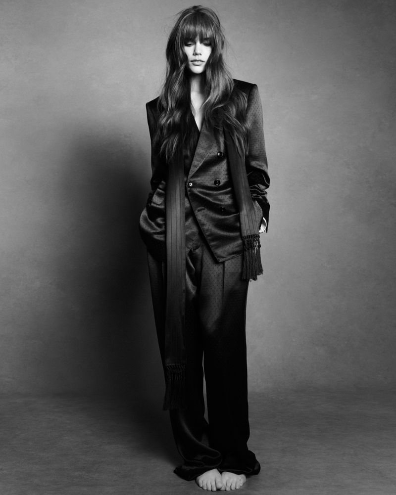 Model wearing a black satin suit with a fringed scarf against a textured grey studio background