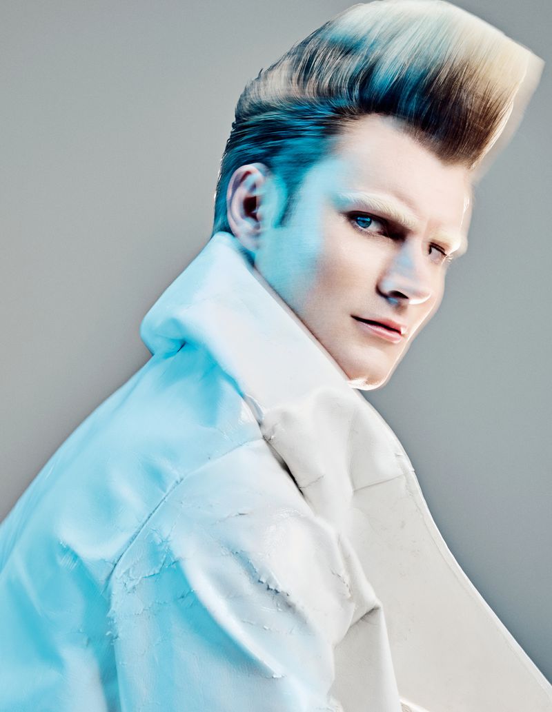 Man with voluminous styled quiff haircut and white jacket looking over his shoulder against a grey background.