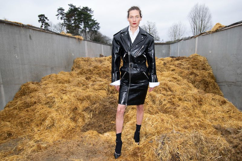 Model wearing a black vinyl coat standing in front of a large pile of straw for Re-Edition Magazine.