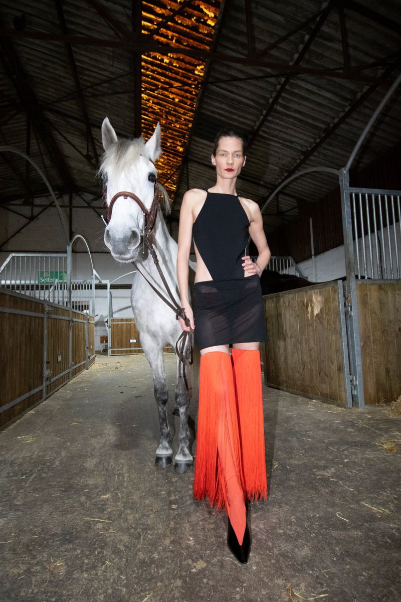 Model wearing a black dress and orange fringed boots standing next to a white horse inside a stable