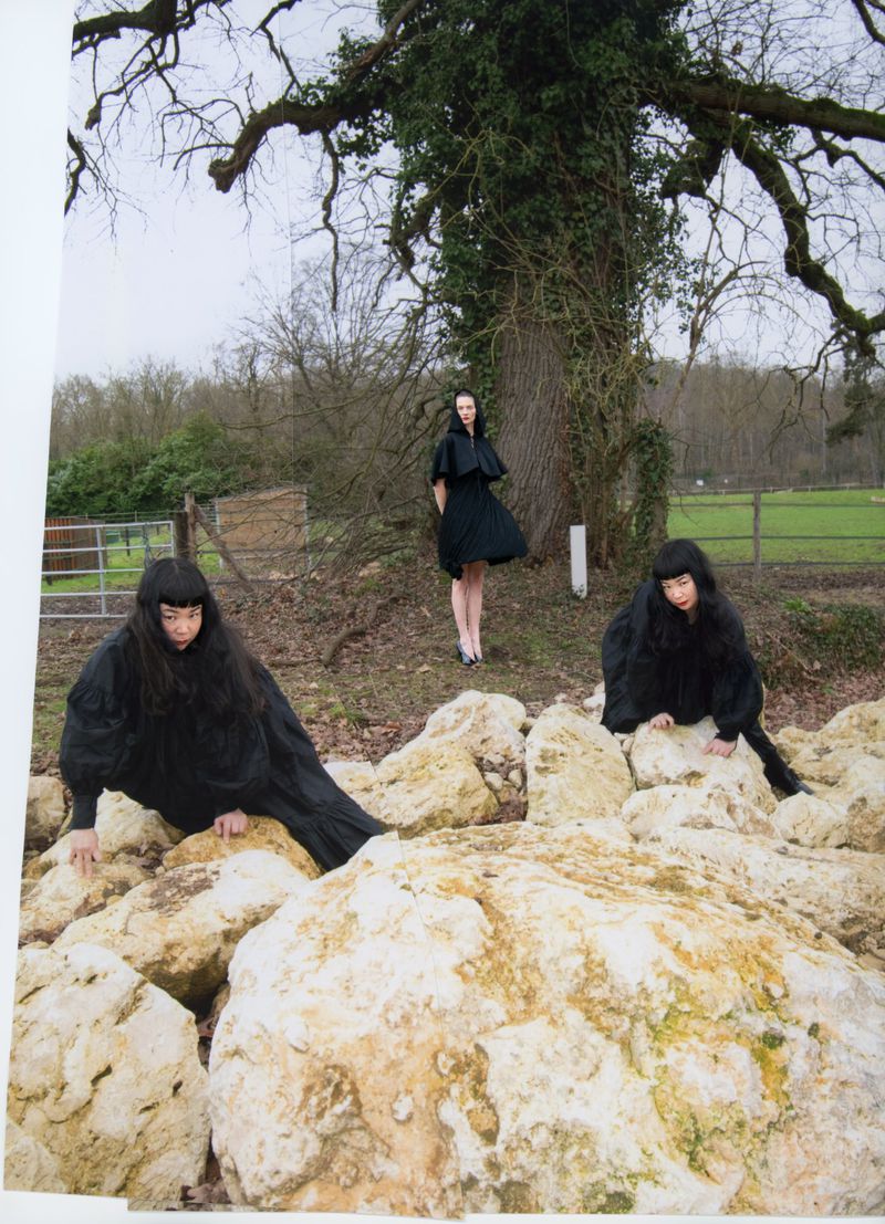 Three models in black dresses posing among large pale boulders outdoors before a large tree and rural fence