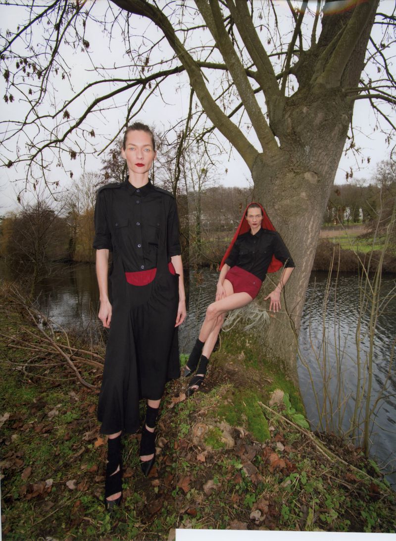 Two fashion models wearing black outfits pose near a large tree and a river. One model is standing, and the other is leaning against the tree.