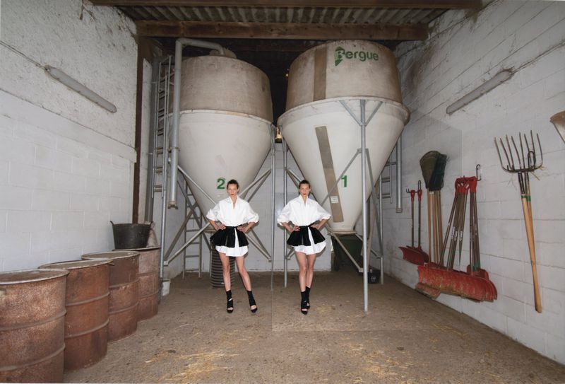 Two models wearing white shirts and dark skirts standing inside an agricultural shed with large silos and farming equipment.
