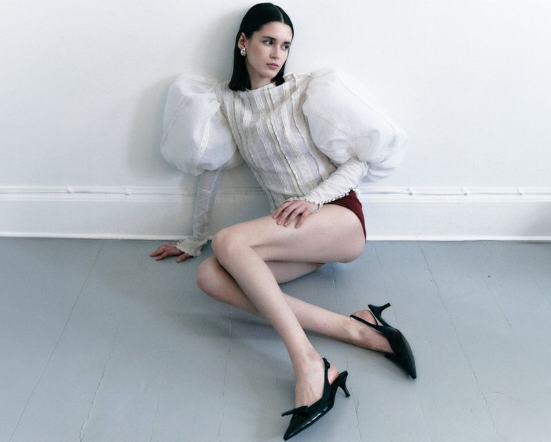 Model with dark hair wearing a white top with voluminous sheer sleeves and black heels seated on a grey floor against a white wall.