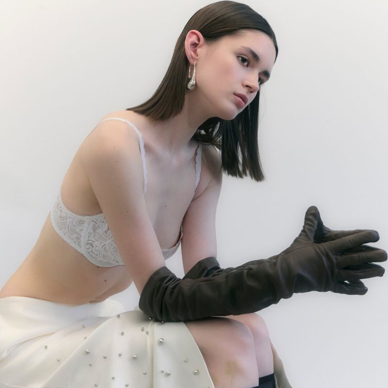 Model in white lace bra and embellished skirt wearing long brown leather gloves against a plain white background.