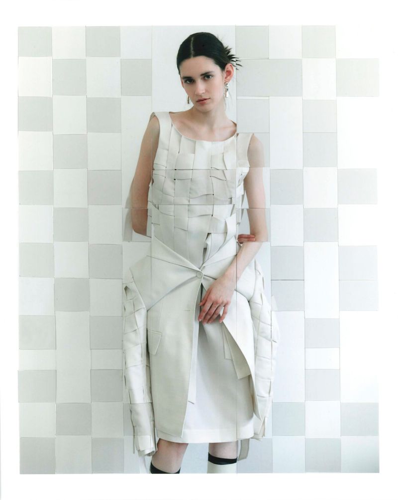Model wearing a structured white woven dress against a pale tiled wall for D'Scene Magazine.