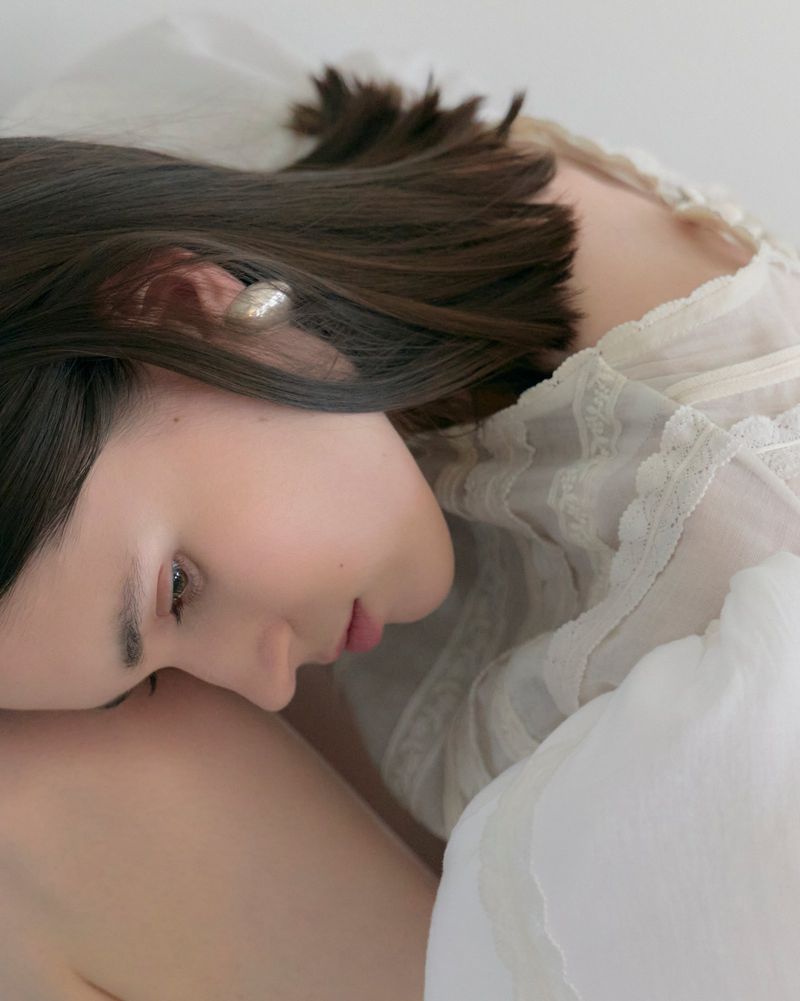 Close-up profile of a woman with dark hair wearing a sheer white blouse and an earring, resting her head against a white pillow.