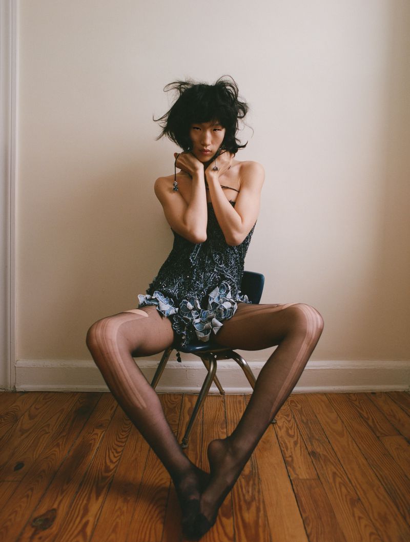Model with dark hair sitting on a small chair against a plain wall wearing a patterned top and sheer dark stockings