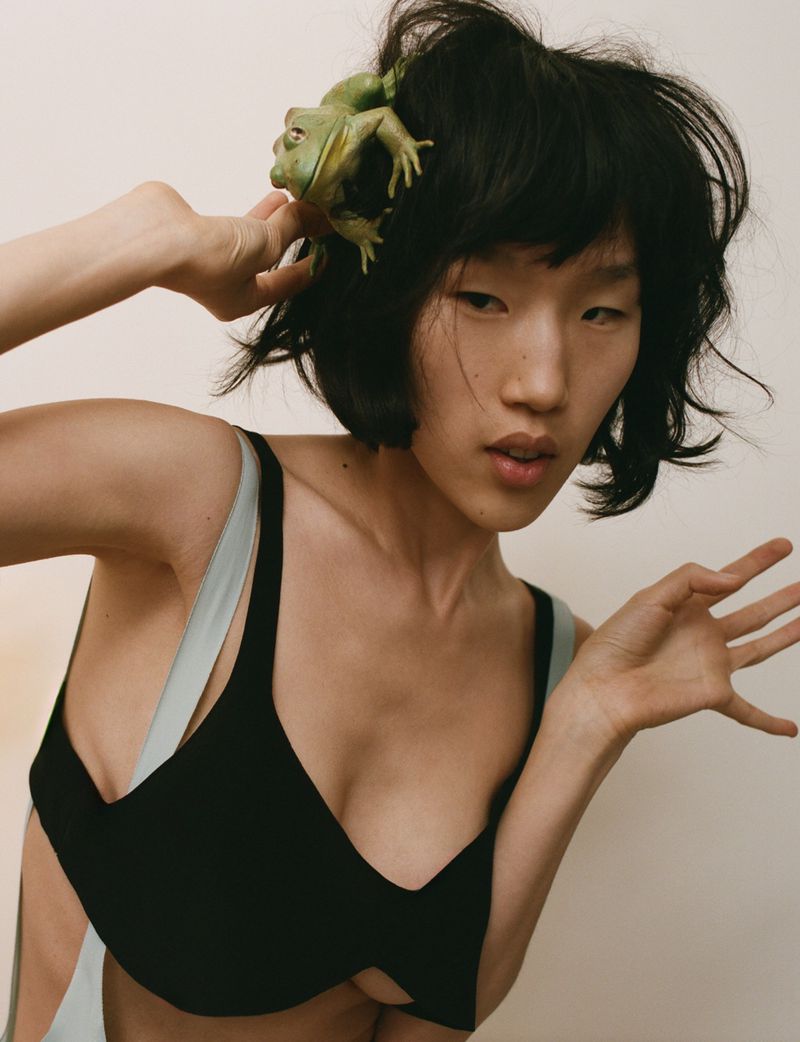 Elegant Asian woman in a black top poses with a green frog perched on her head, set against a minimalist backdrop.