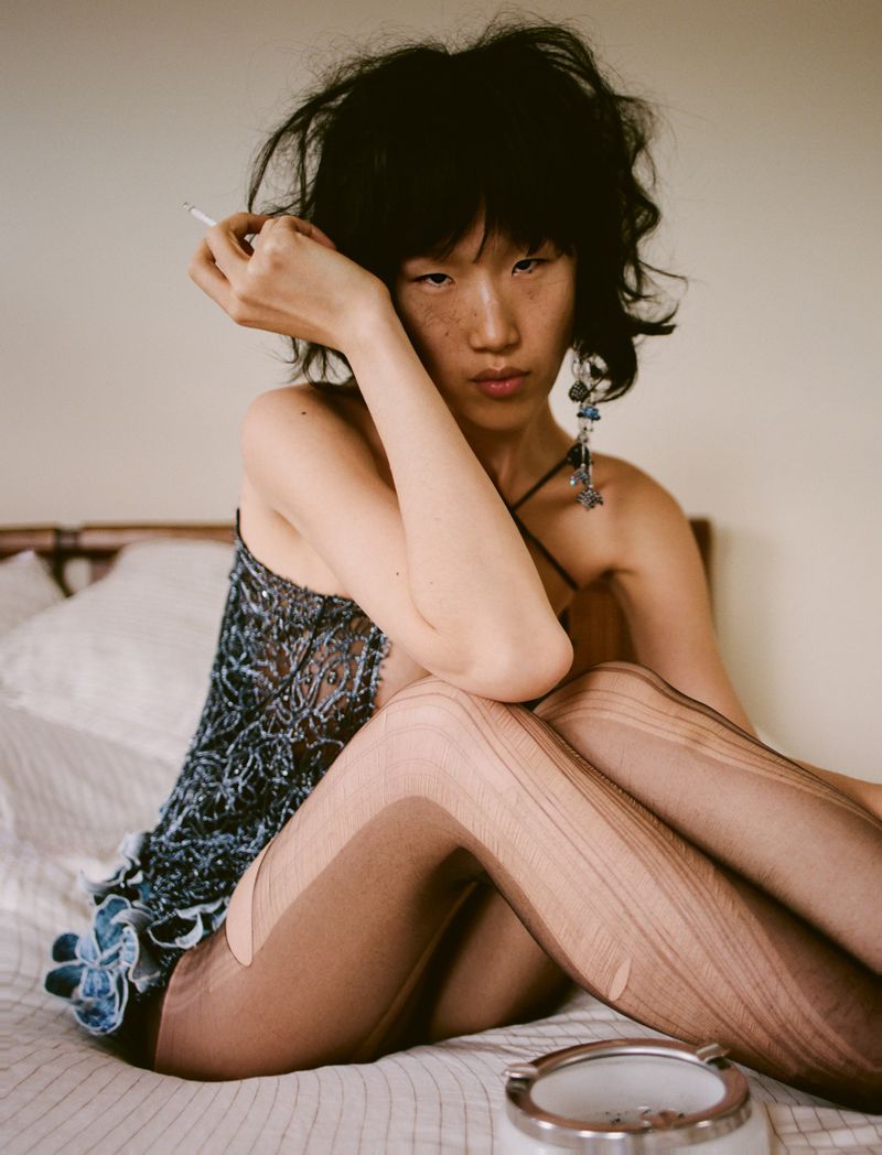 Model with dark tousled hair wearing a patterned top and patterned tights, seated on a bed with a white pillow and ashtray.