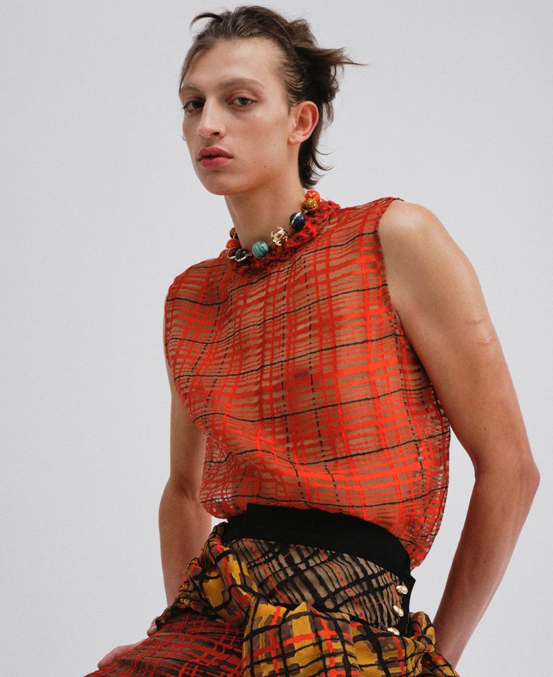 Model wearing a red patterned sheer sleeveless top and beaded necklace against a plain white background