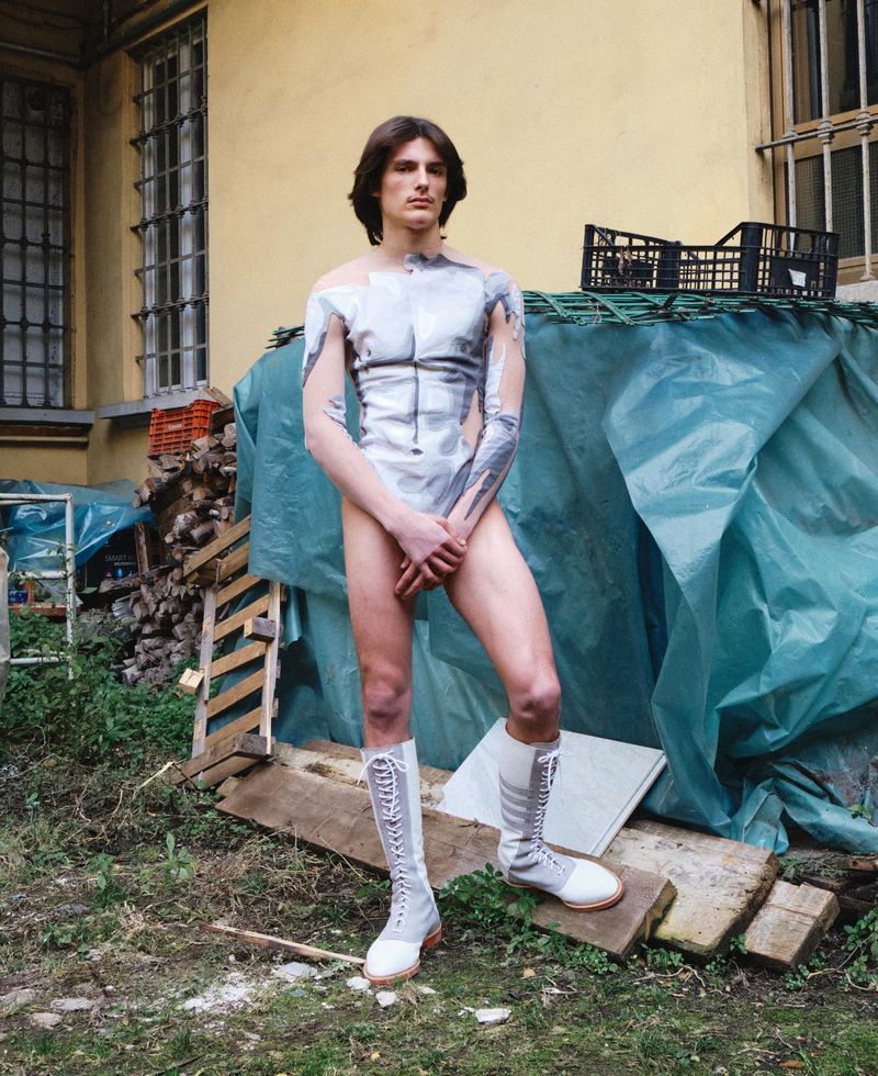 Model standing outdoors wearing knee-high lace-up boots and a printed body-hugging top before a blue tarp and yellow building.