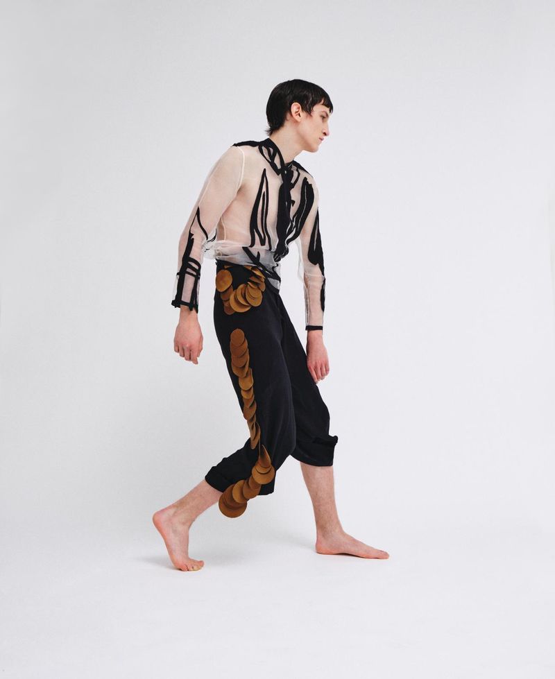 Model walking barefoot wearing a sheer patterned shirt and black trousers decorated with circular patches for ODDA Magazine