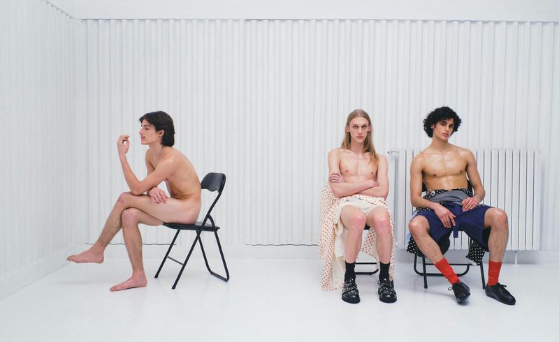 Three male models seated on black folding chairs against a vertical white paneled background for ODDA Magazine