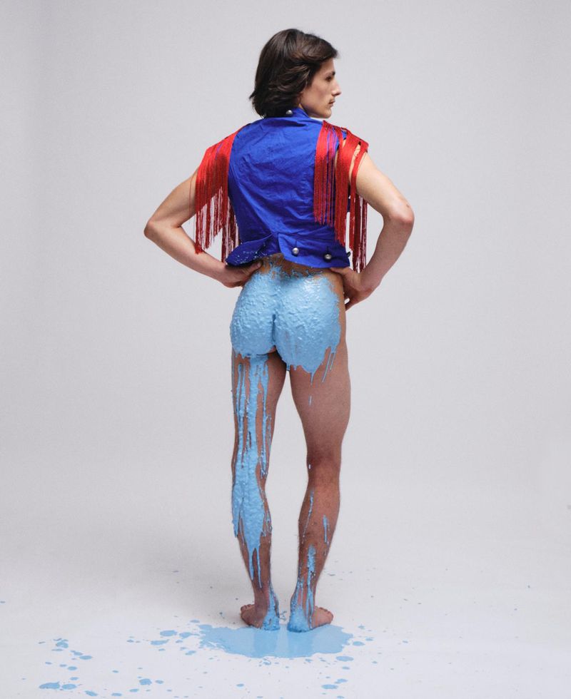 Model from behind wearing a blue vest with red fringe and blue paint dripping down his legs against a white background