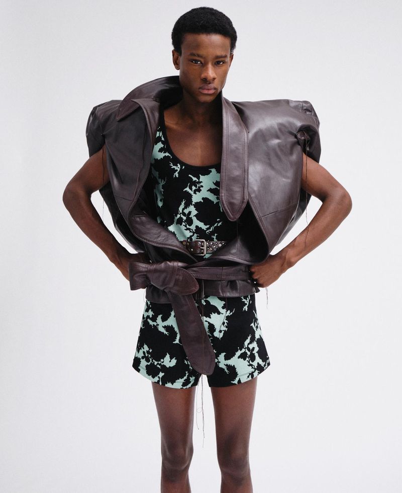 Model wearing an oversized brown leather jacket and patterned shorts against a white background