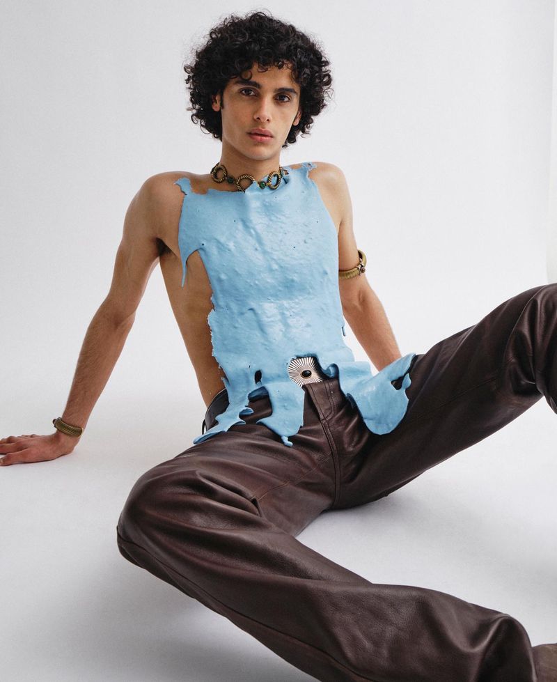 A young male model with dark curly hair seated on a white floor wearing a textured light blue top and dark brown trousers.