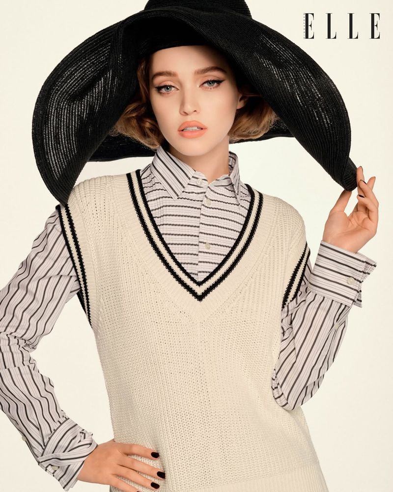 Model Polly Domashych wearing a large black sun hat, striped shirt, and cream sweater vest against a white seamless backdrop.