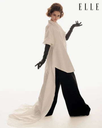 Model wearing a white tunic, black trousers, and long dark gloves against a plain white background in an editorial fashion shoot