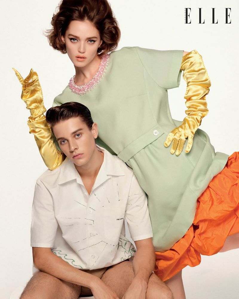 Model standing behind a seated man, wearing a mint green dress with yellow satin gloves against a white background